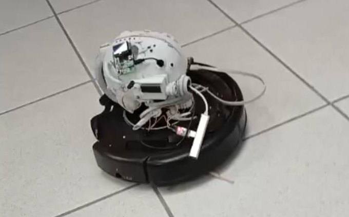 Roomba preto com um Raspberry Pi e uma câmera parafusados por cima, fiação aparente, num piso de cerâmica